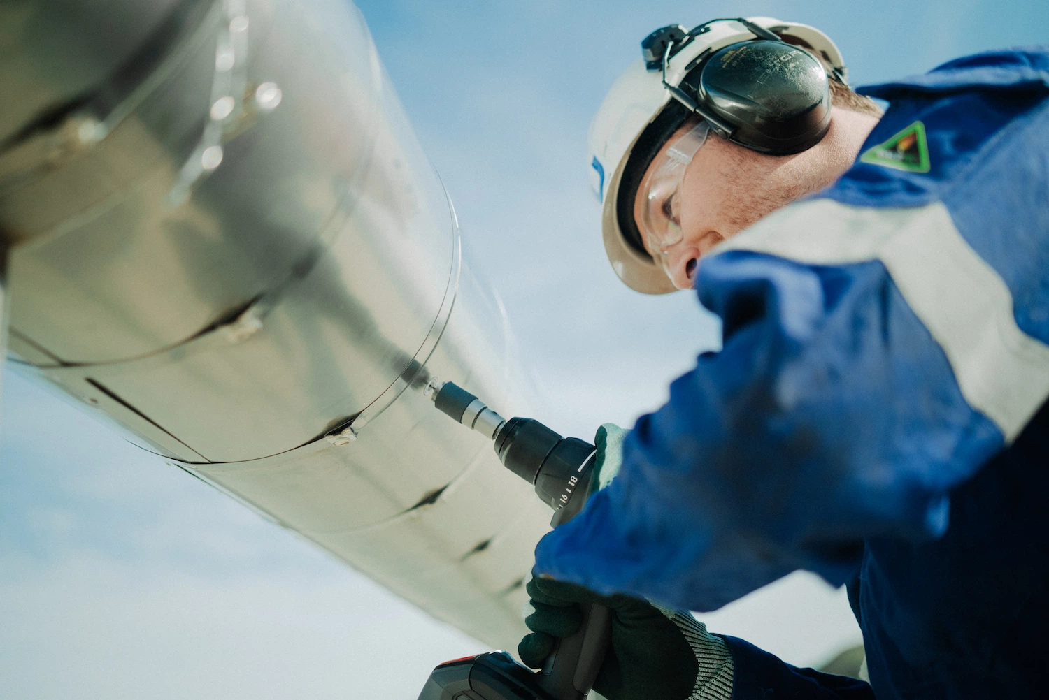 Technician in helmet and PPE drilling into an insulated industrial vessel during inspection